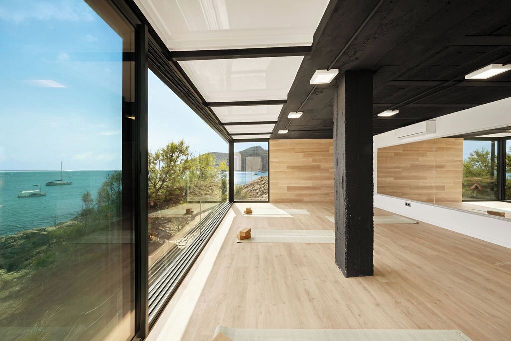 A spacious room with large windows overlooking a body of water and a view of boats. The room features a wooden floor and a glass partition.