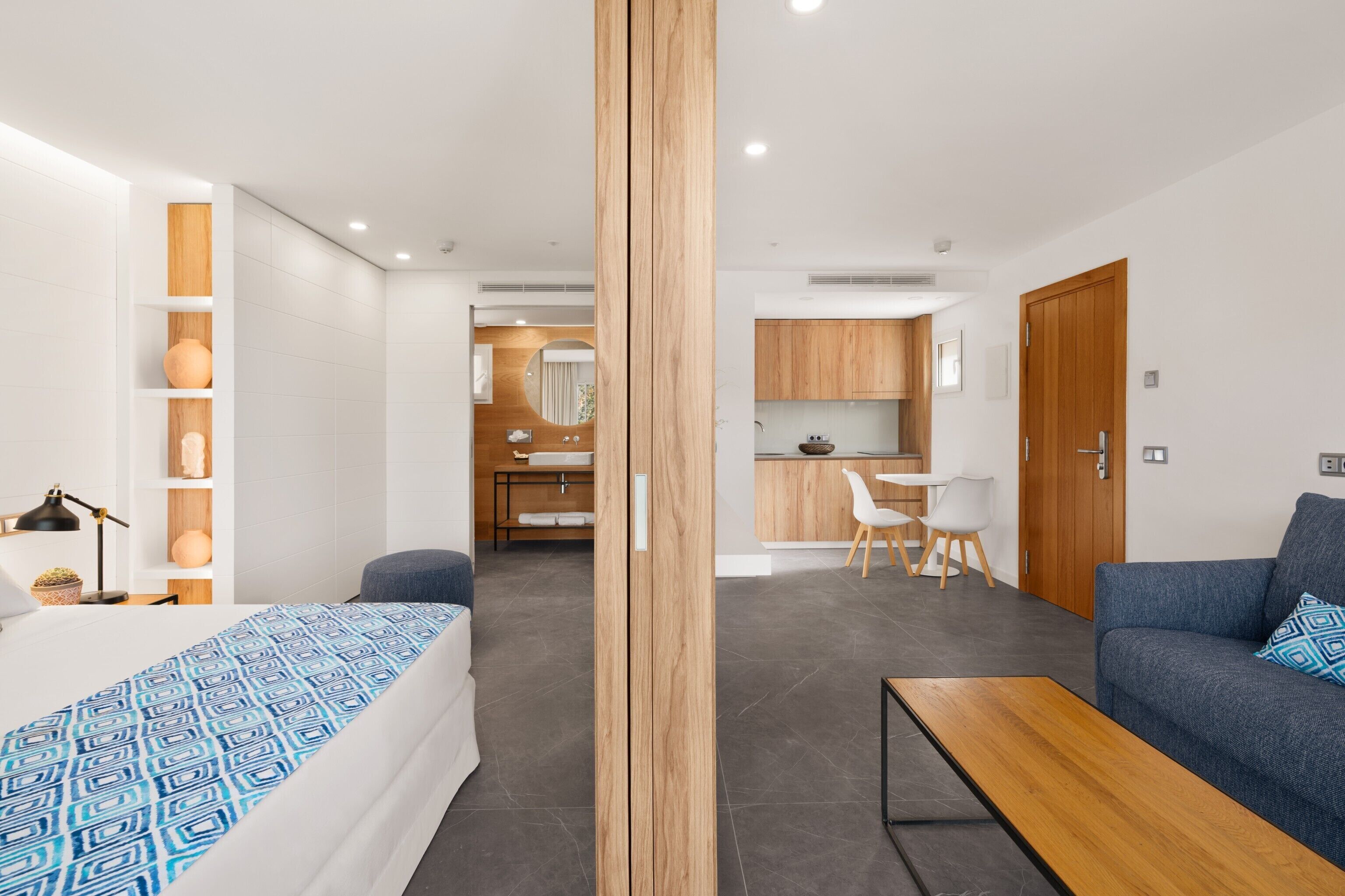 A modern hotel room with a bed, a wooden partition, a sofa, a dining area, and a kitchen.