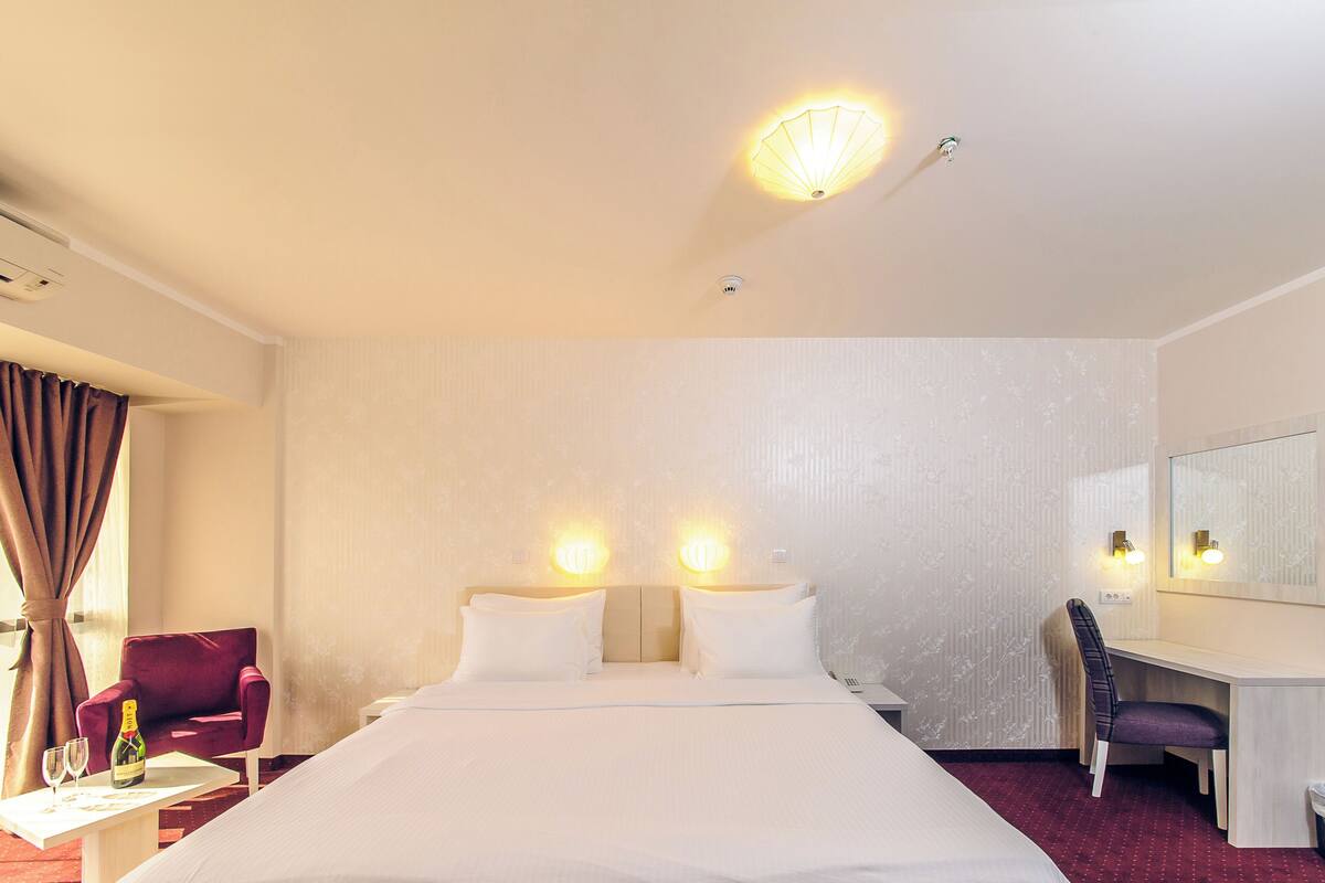 A hotel room with a bed, two bedside lamps, a desk with a chair, a mirror, and a trash bin.
