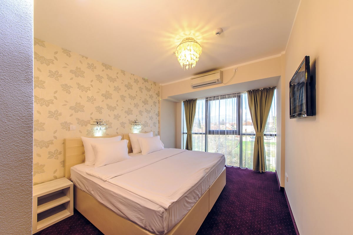 A hotel room with a bed, a nightstand, a window with curtains, an air conditioning unit, and a mounted TV.