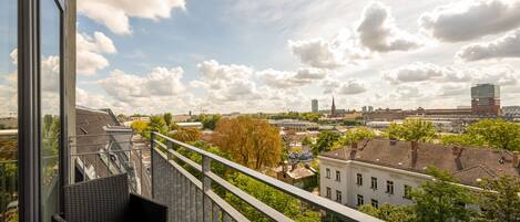 Un balcón con una silla de mimbre y una mesa, con vistas a un paisaje urbano con edificios y árboles.