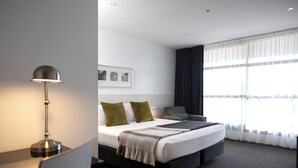 A modern hotel room with a large bed, a desk, and a chair. There is a window with sheer curtains and a view of the city. - Quest Parnell (Auckland)