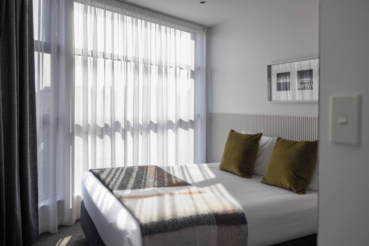 A hotel room with a bed, a window with sheer curtains, a framed picture on the wall, and a radiator.