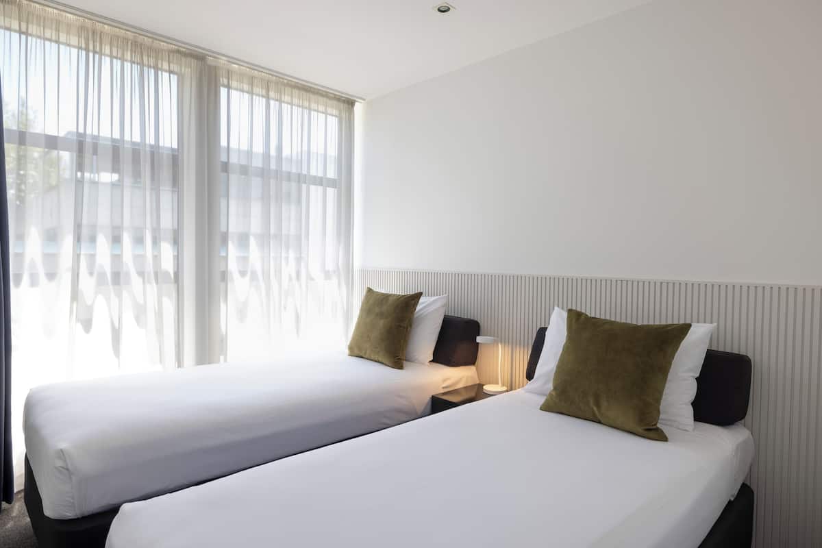 A hotel room with two beds, a nightstand with a lamp, and a window with sheer curtains.