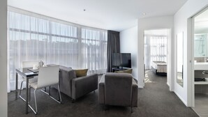 A modern hotel room with a sofa, armchair, and a dining table with chairs. There is a TV and a bathroom visible through an open door. - Quest Parnell (Auckland)
