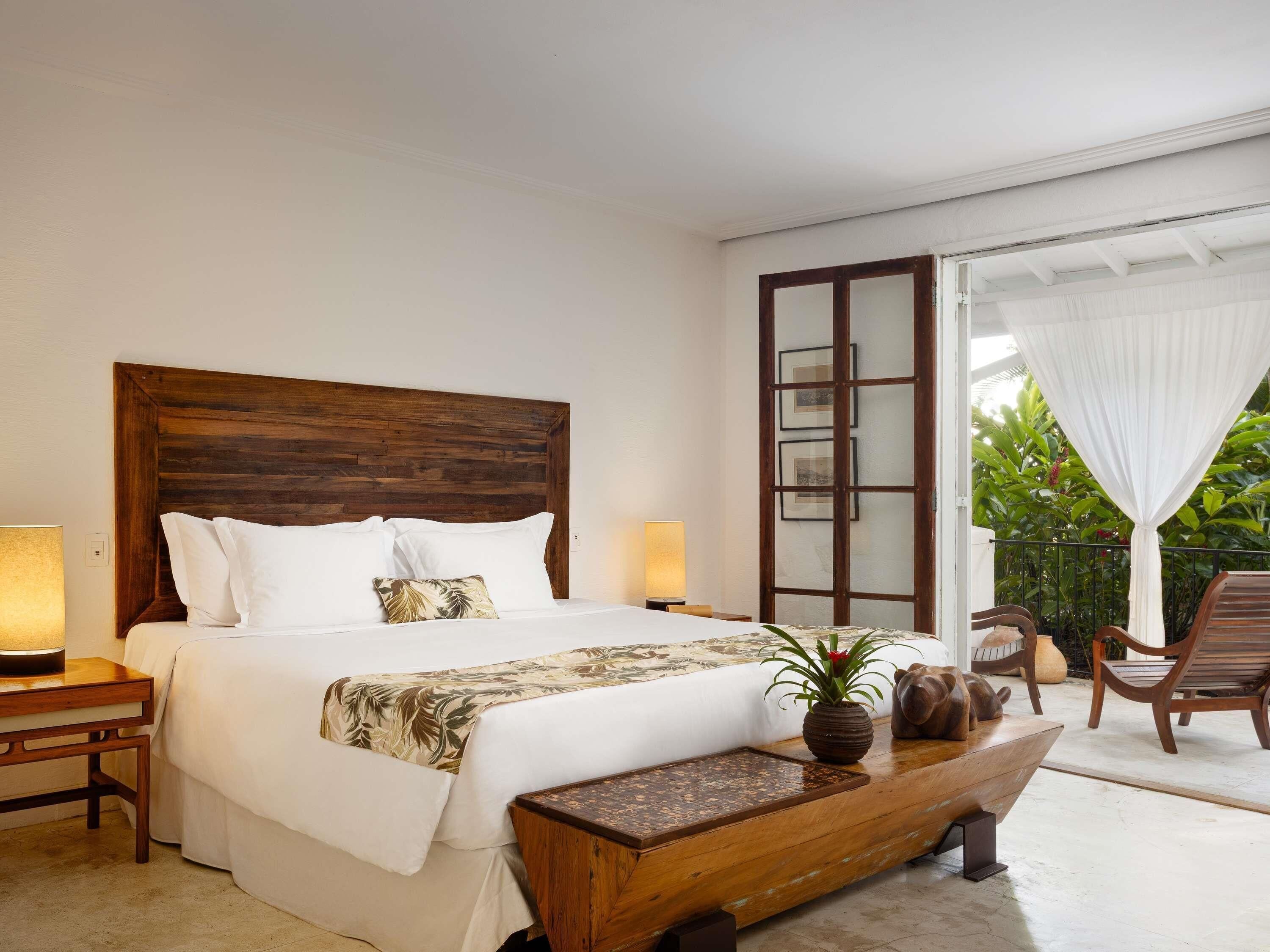 A bedroom with a wooden headboard, a bed with white linens, a bedside table with a lamp, and a view of the outdoors through a glass door.