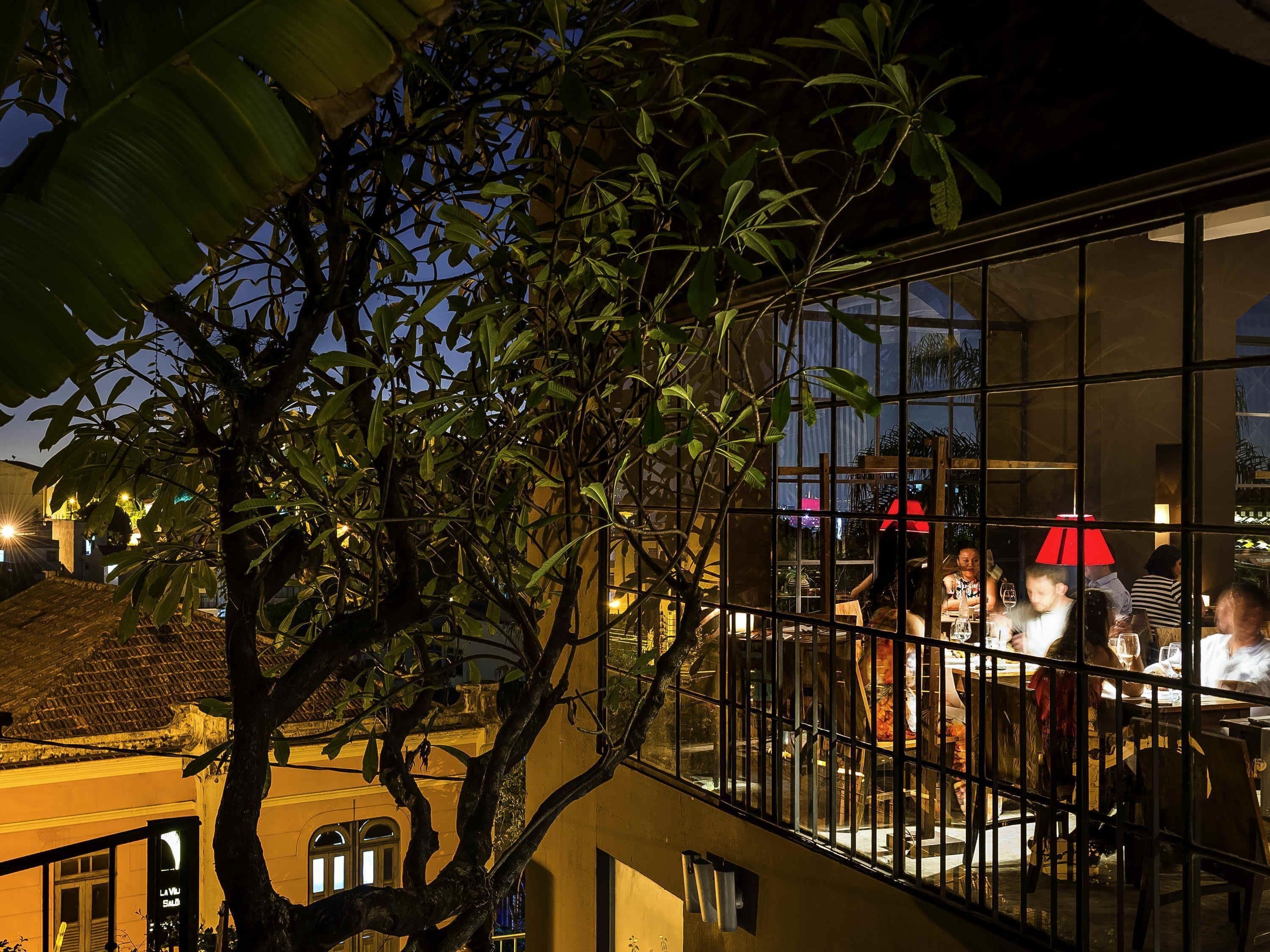 Una terraza con comedor, rodeada de abundante vegetación y un edificio con amplios ventanales.