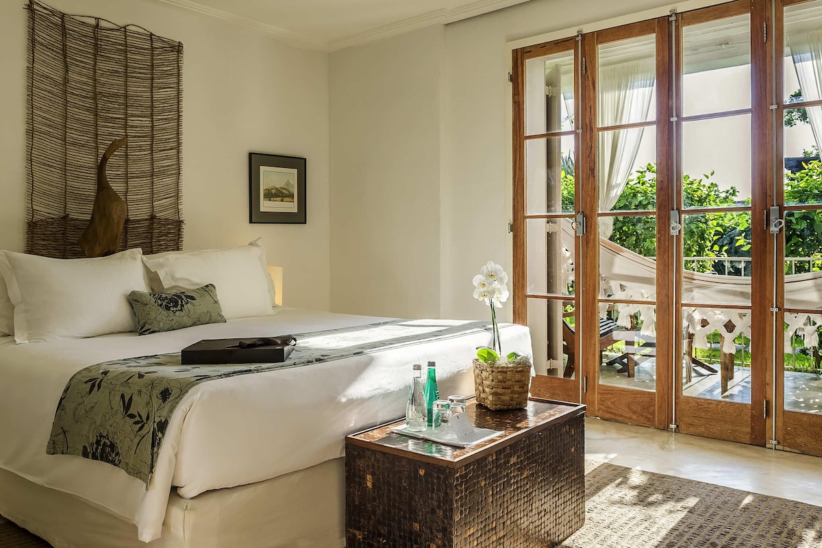 A bedroom with a bed, a bedside table with a vase, a woven wall hanging, and a door leading to an outdoor area.