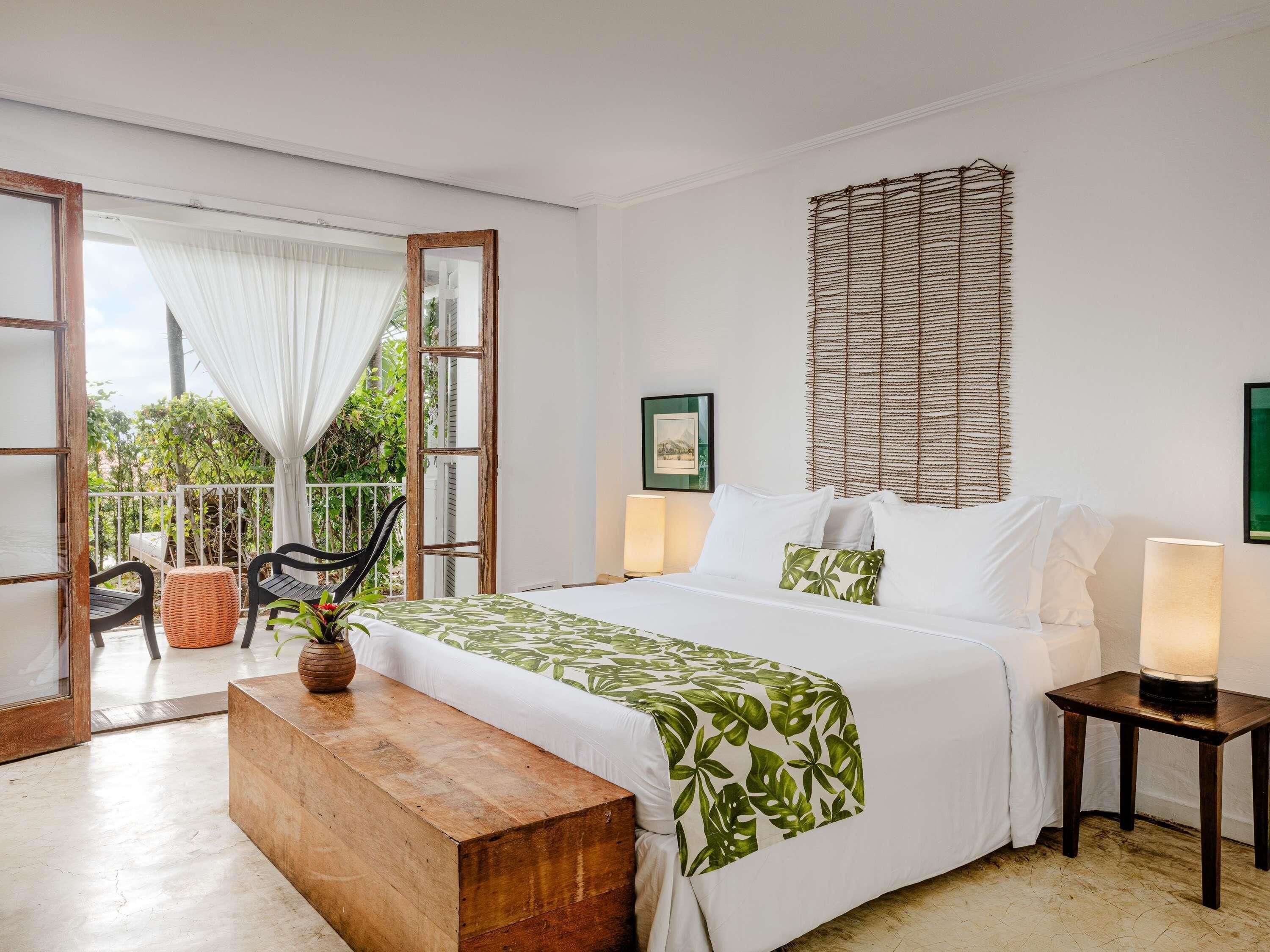 A bedroom with a bed, bedside lamps, a wooden bench, a wicker headboard, and a view of the outdoors.