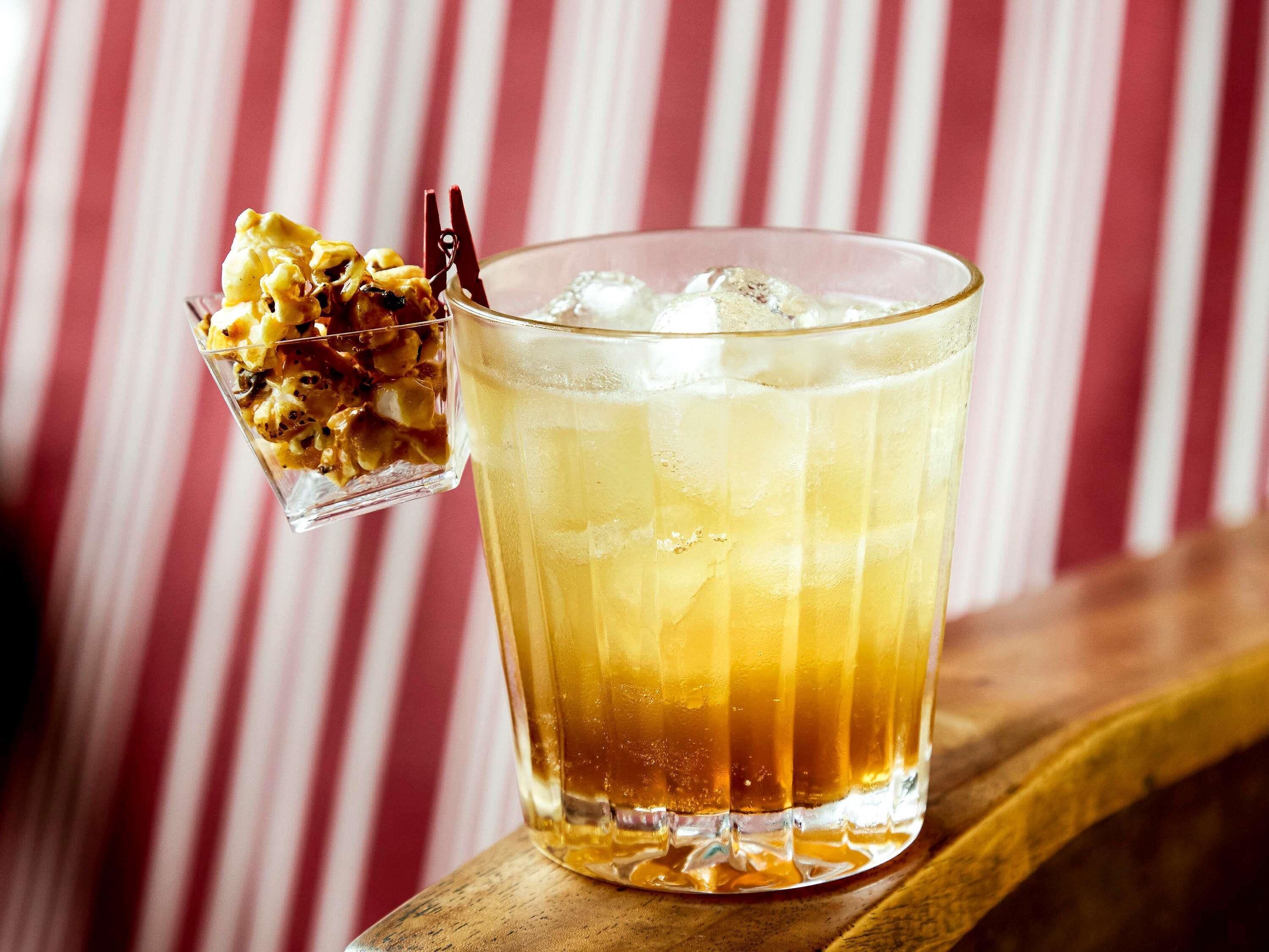 A glass of iced beverage with a fruit garnish on the side, placed on a wooden surface with a striped background.