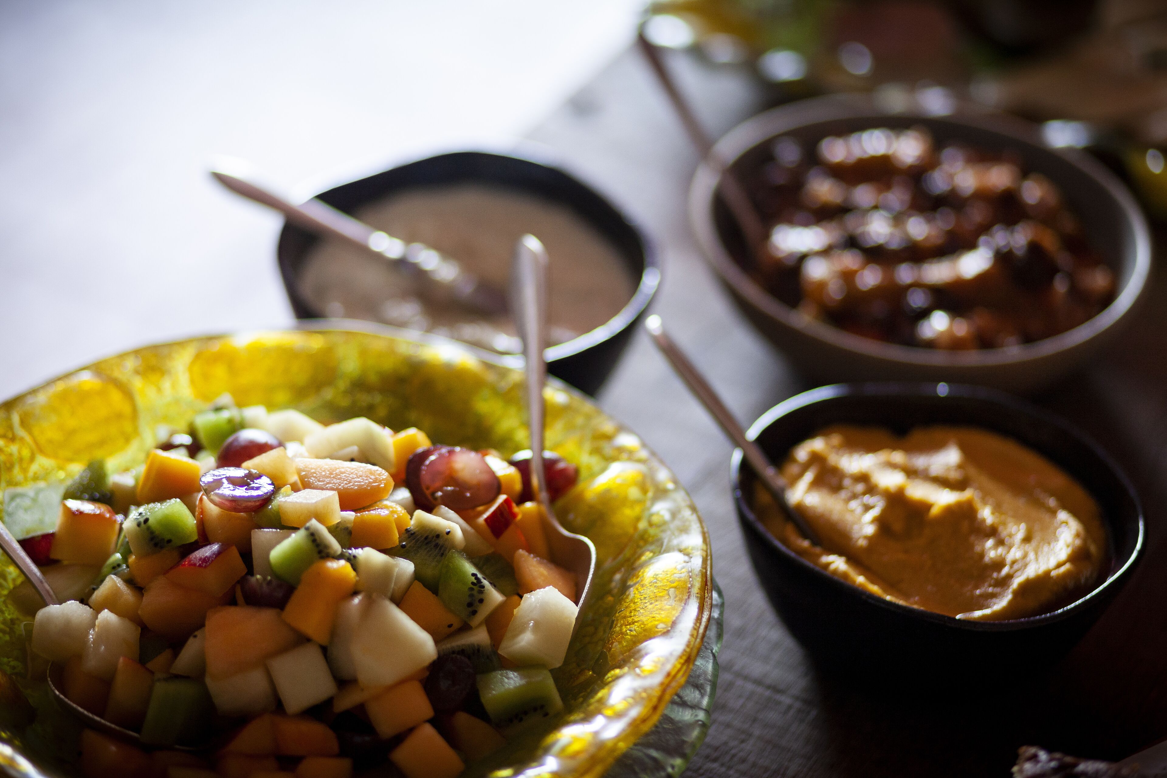 A variety of food items including a fruit salad, a bowl of soup, and a bowl of hummus.