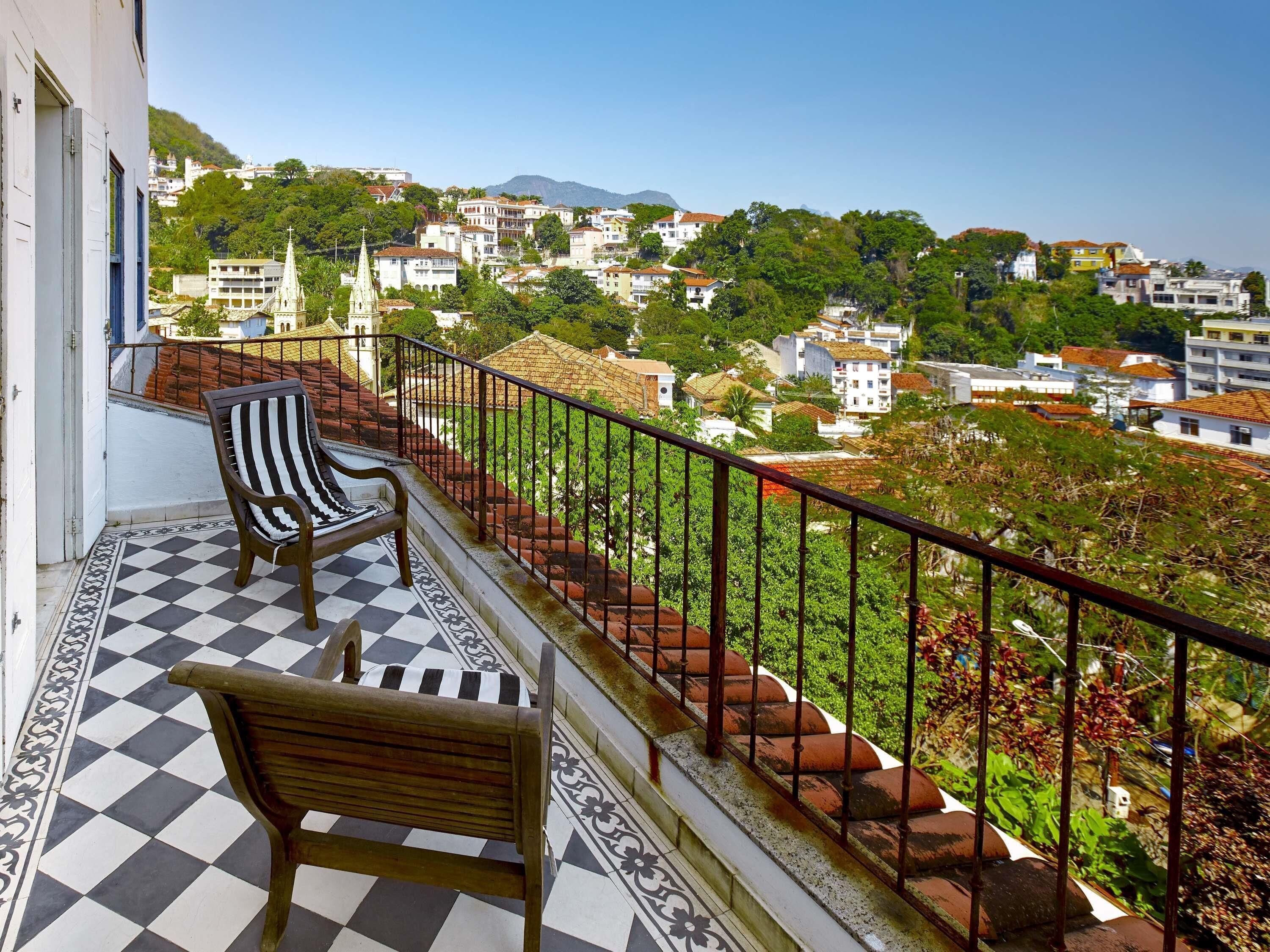 A balcony with a checkered floor, two wooden chairs, and a metal railing overlooking a hillside with buildings.