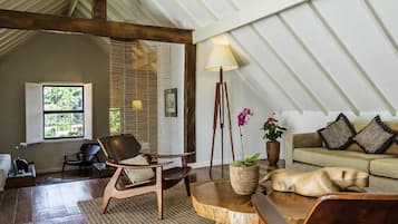 A cozy loft-style room with a high ceiling, a wooden floor, a round coffee table, a leather armchair, a sofa with cushions, a floor lamp, and a window with shutters.