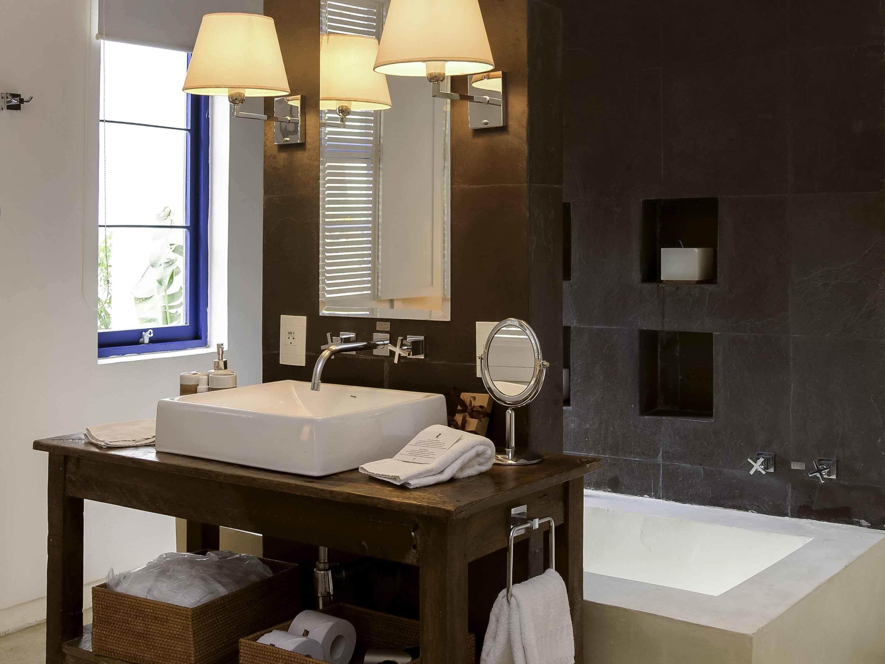Un baño con un lavamanos grande de color blanco, un espejo y una bañera. Hay tres lámparas empotradas en la pared sobre el lavamanos.