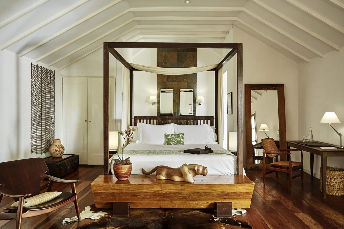 A four-poster bed with a dog, a wooden desk, a chair, and a mirror in a room with wooden flooring and white walls.
