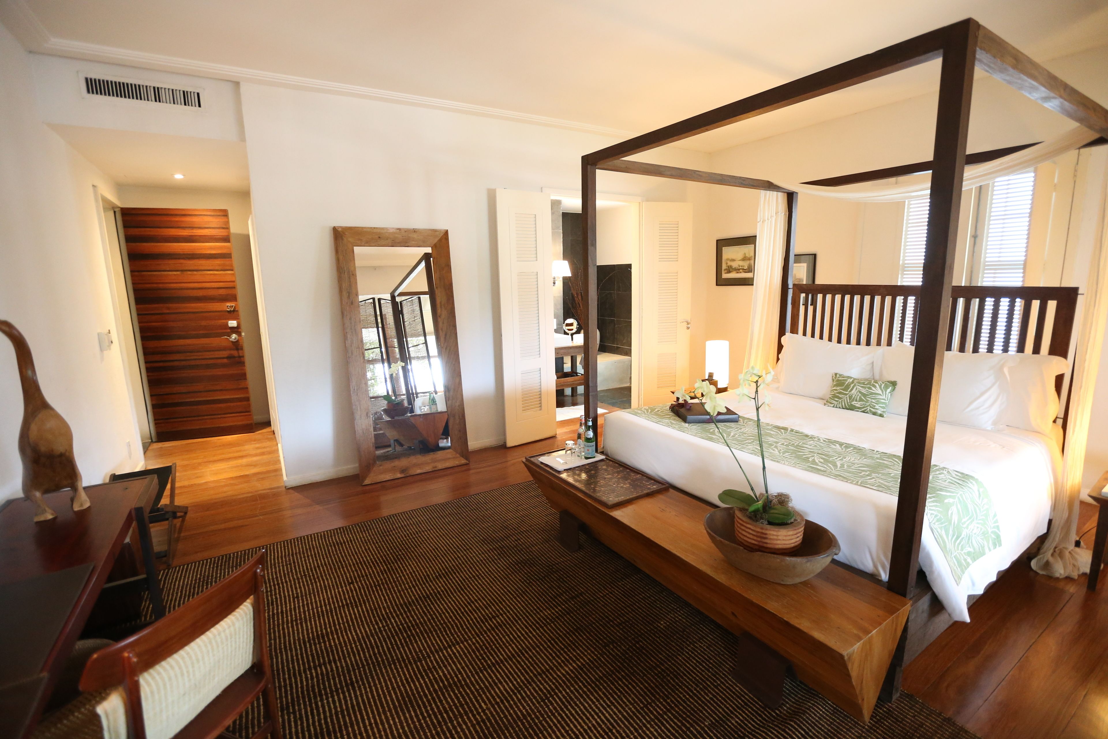 A bedroom with a four-poster bed, a wooden bench, a dining table with chairs, and a large mirror.
