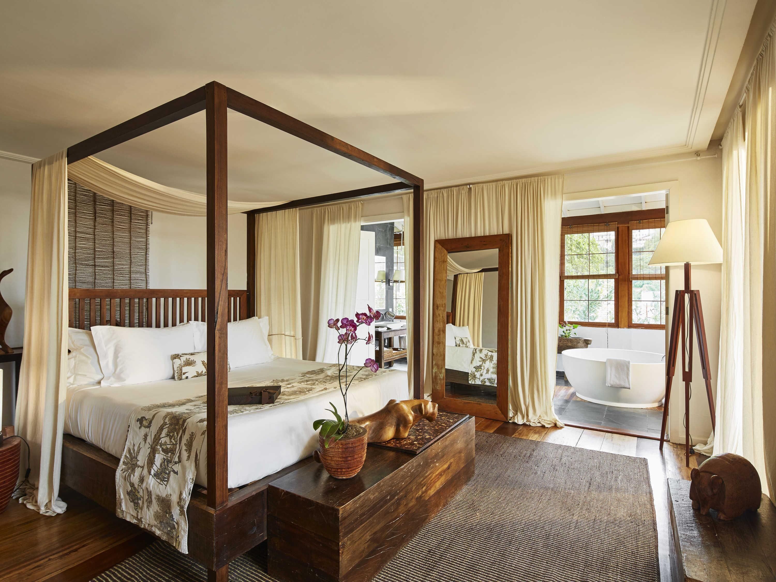 Un lit à baldaquin avec une tête de lit en bois, une table de chevet agrémentée d’une plante, et une vue sur une salle de bain avec une baignoire.