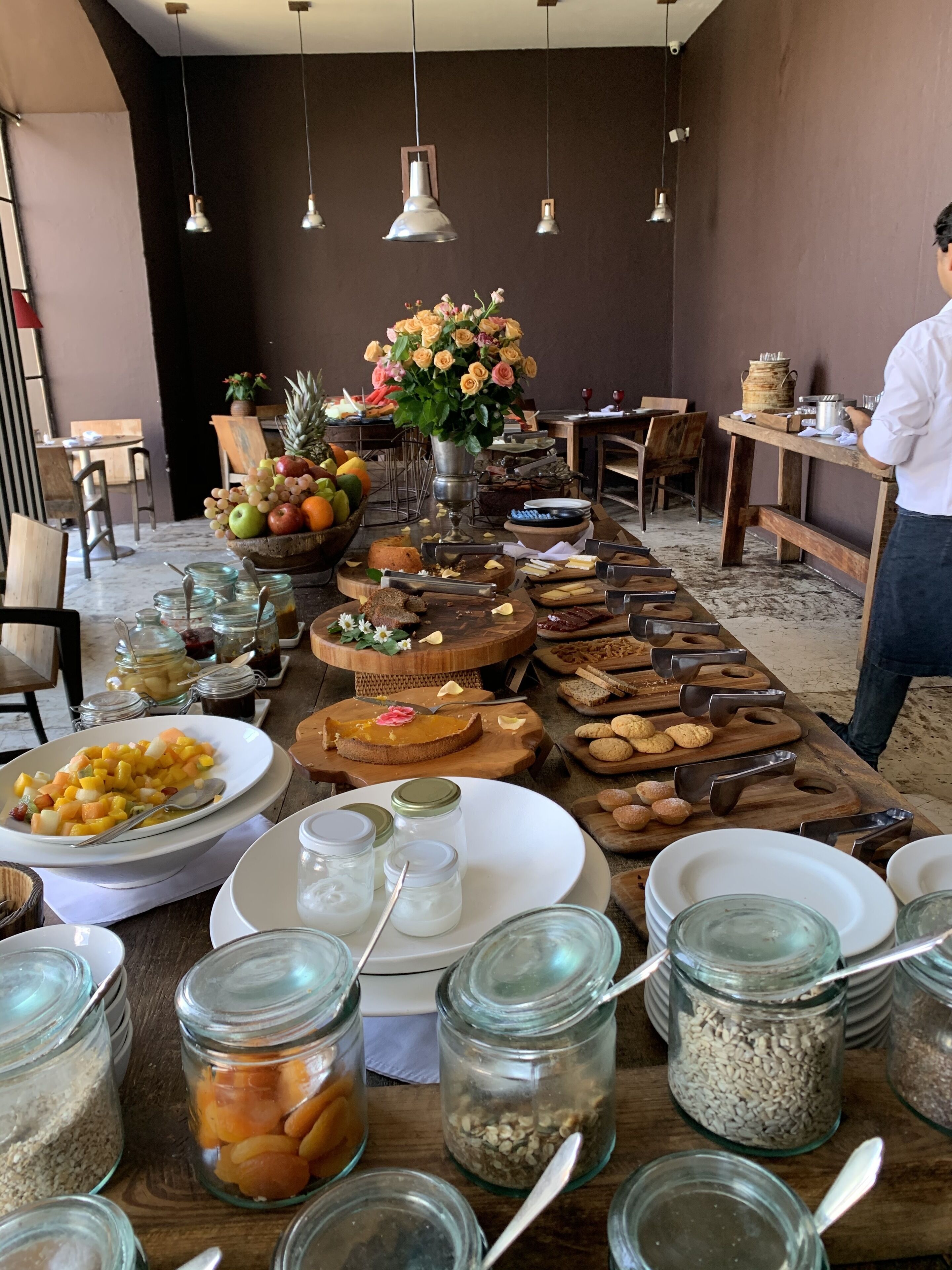 Buffet com diversos alimentos e utensílios, incluindo frutas, doces e condimentos em potes de vidro.