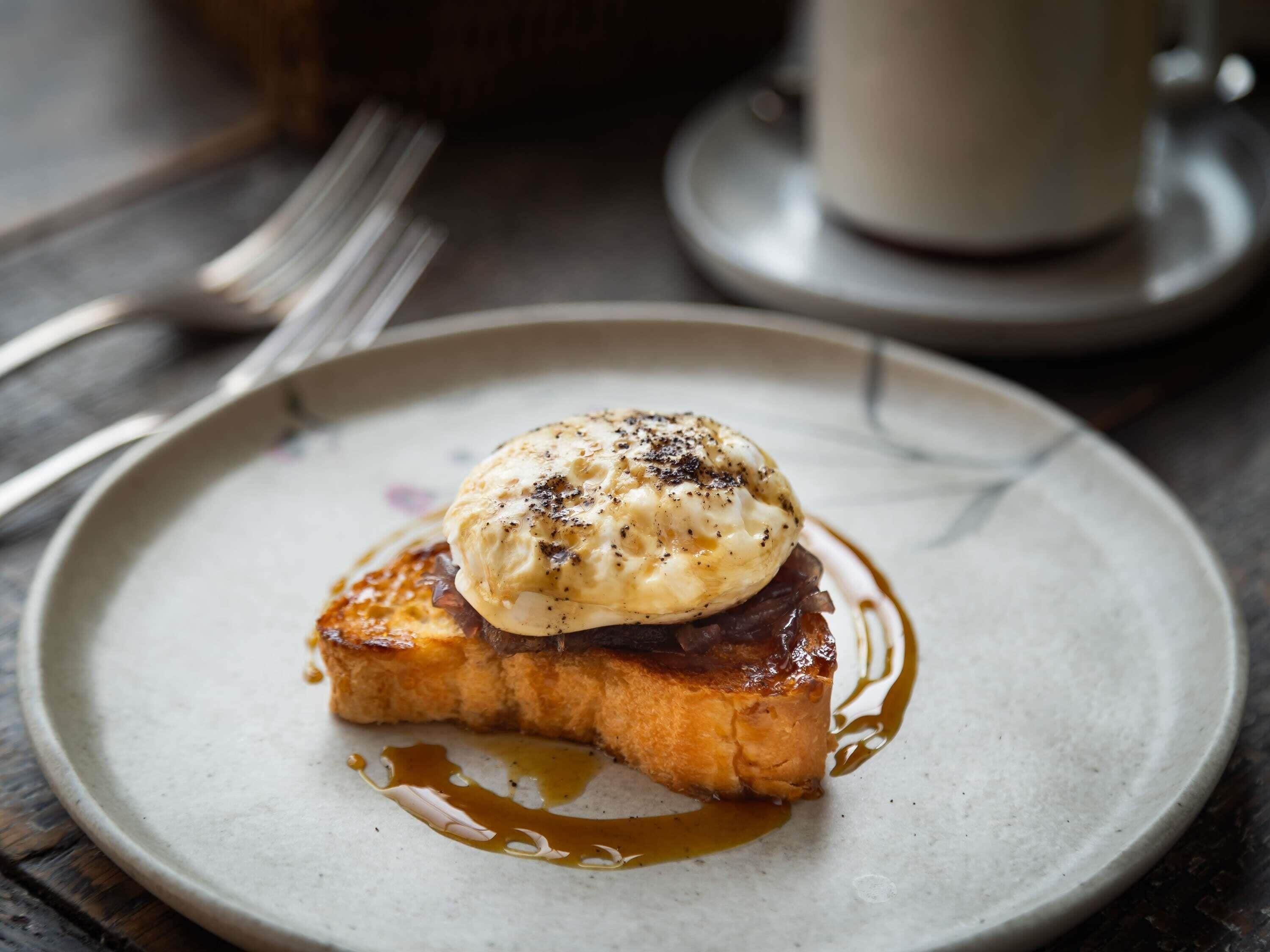 A plate with a food item, possibly a sandwich or pastry, topped with a creamy sauce and a poached egg, accompanied by a cup and saucer in the background.
