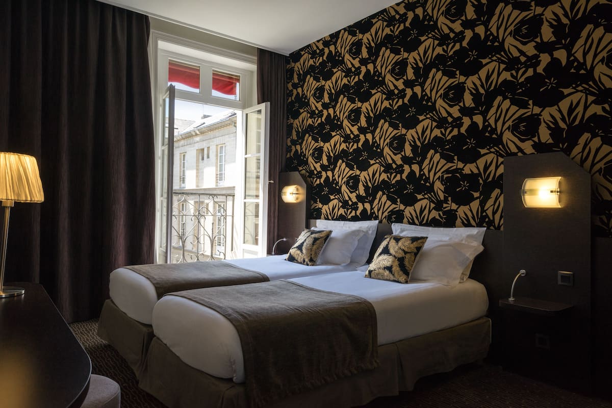 Een hotelkamer met twee bedden, een raam met gordijnen, een behang met patroon en een lampje op het nachtkastje.