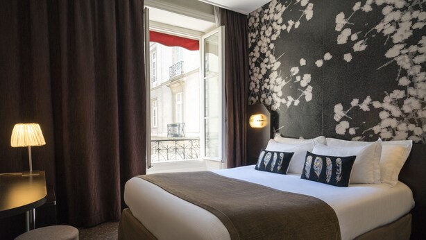 A hotel room with a large bed, a desk with a lamp, a view of the outside balcony, and a floral-patterned wall. - Hôtel Voltaire Opéra (Nantes)