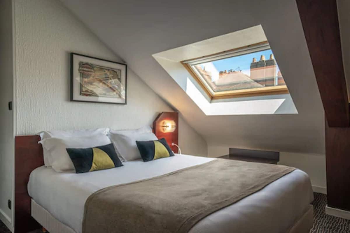 Slaapkamer met dakraam, een bed met kussens, een ingelijst schilderij aan de muur en een nachtkastje met een lamp.
