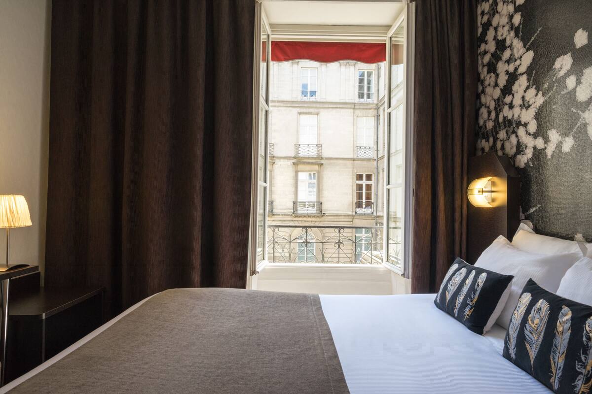 Hotelkamer met groot bed, bedlamp, uitzicht op een gebouw door een raam en decoratieve wandversiering.