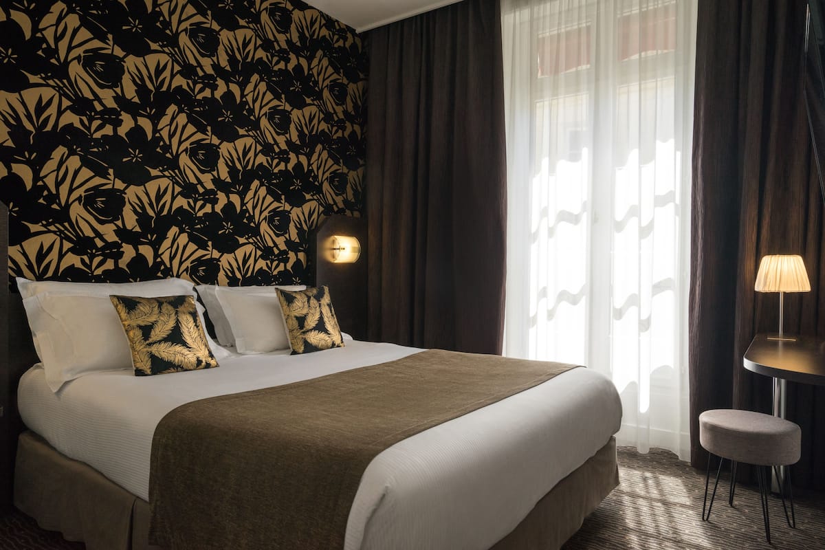 Hotelkamer met een groot bed, donkere gordijnen, een wandlamp en een tafeltje met een lamp.
