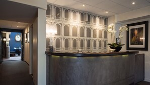 A reception area with a curved reception desk, a large ornate wall with arches, a clock, and a framed picture. - Hôtel Voltaire Opéra (Nantes)