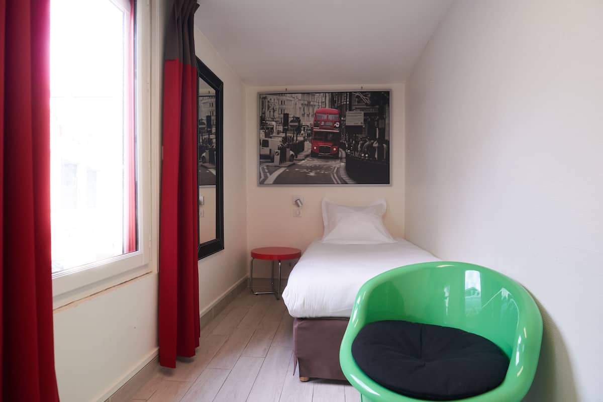 A bedroom with a bed, a green chair, a red table, and a cityscape painting on the wall.