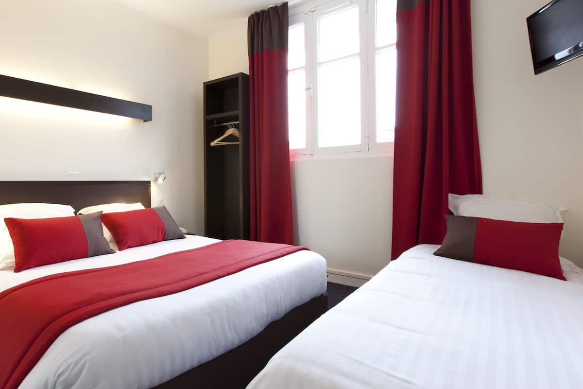 A hotel room with two beds, red and white bedding, a dark headboard, a wall-mounted TV, and a window with red curtains.
