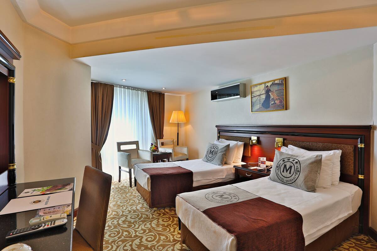 A hotel room with two beds, a desk with a lamp, a chair, and a TV on the wall.