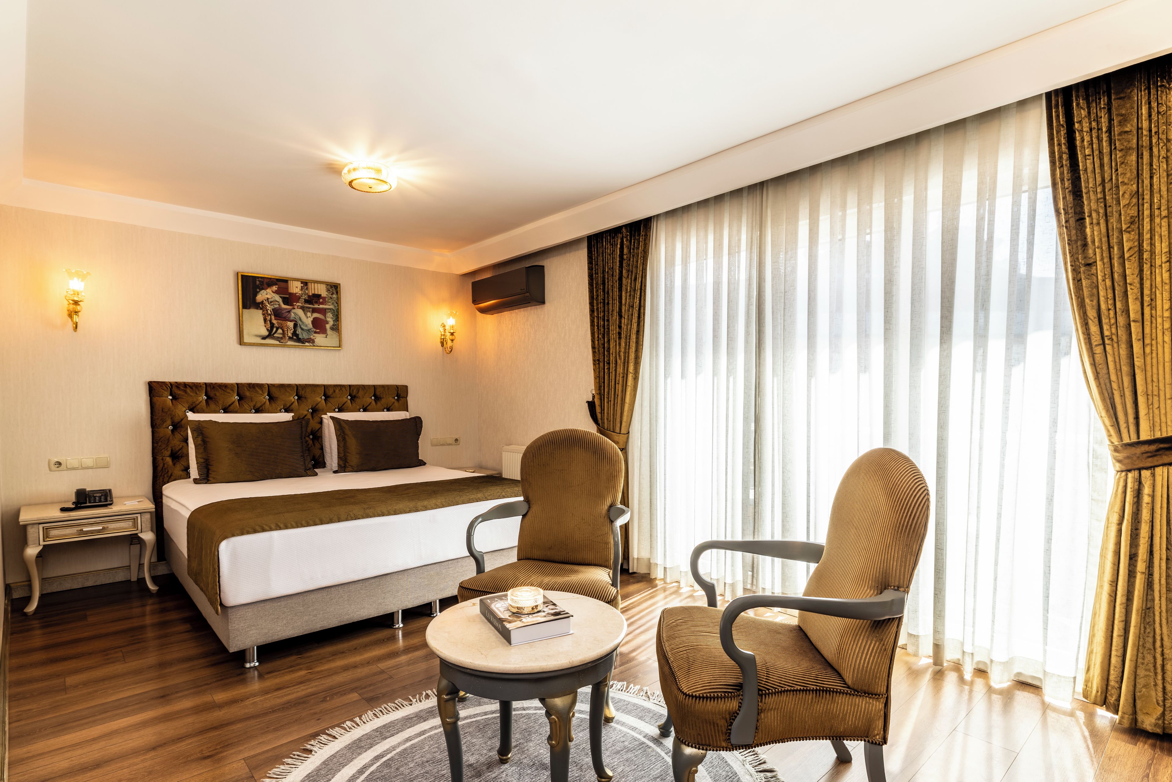 Habitación de hotel con cama, dos sillas, una mesita, un cuadro, un televisor instalado en la pared y una ventana con cortinas.