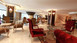Un hall espacioso con diversas opciones de asientos, incluyendo sillones de terciopelo rojo y sillones tapizados en marrón, una mesa de centro de vidrio y una gran lámpara de araña.