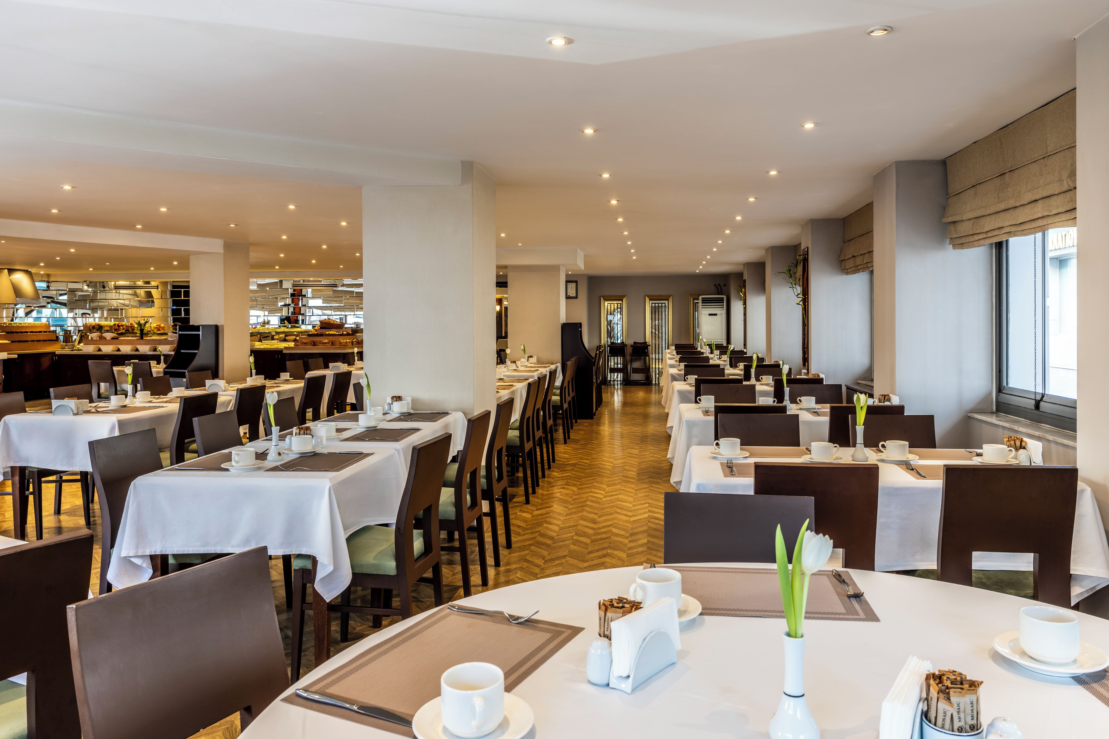 Une salle à manger d’hôtel avec de nombreuses tables préparées pour le petit-déjeuner, une cuisine en arrière-plan et une vue sur la ville à travers de grandes fenêtres.