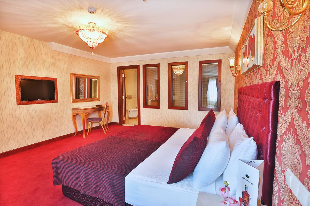 A hotel room with a large bed, a desk, a chair, a television, and a chandelier.