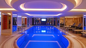 Une piscine intérieure moderne, dotée d’un sol illuminé de lumière bleue, entourée de chaises longues et agrémentée d’un mur miroir.