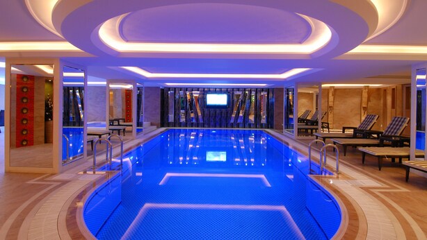 Una piscina interior moderna con suelo iluminado en azul, rodeada de sillones de descanso y un muro espejado.