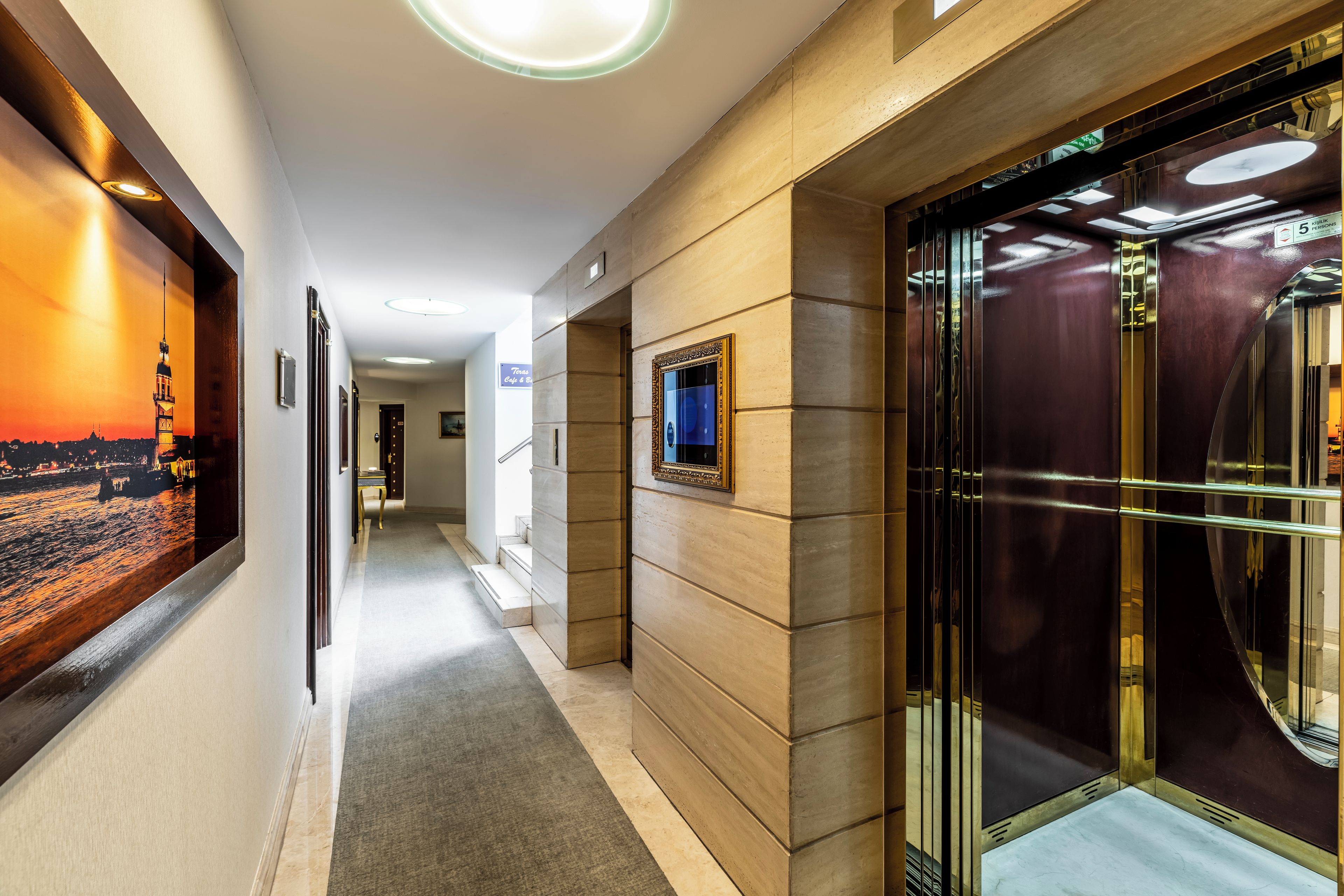 A hotel corridor with a framed picture of a cityscape, a sign for a cafe, and an elevator with a glass door.