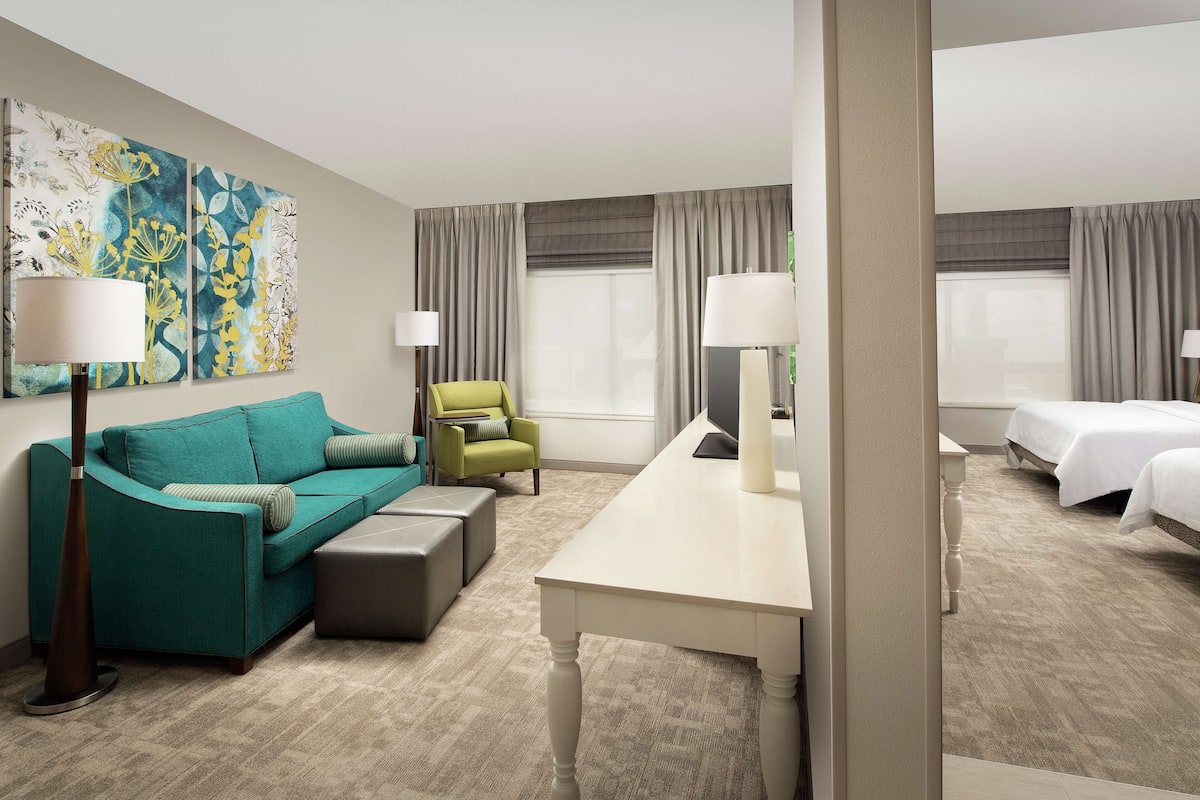 Une chambre d’hôtel avec un canapé turquoise, un fauteuil vert, une table blanche et deux lits.