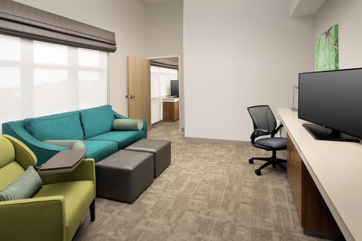 Une chambre d’hôtel moderne avec un canapé vert, un fauteuil vert, un bureau avec un ordinateur et une télévision.