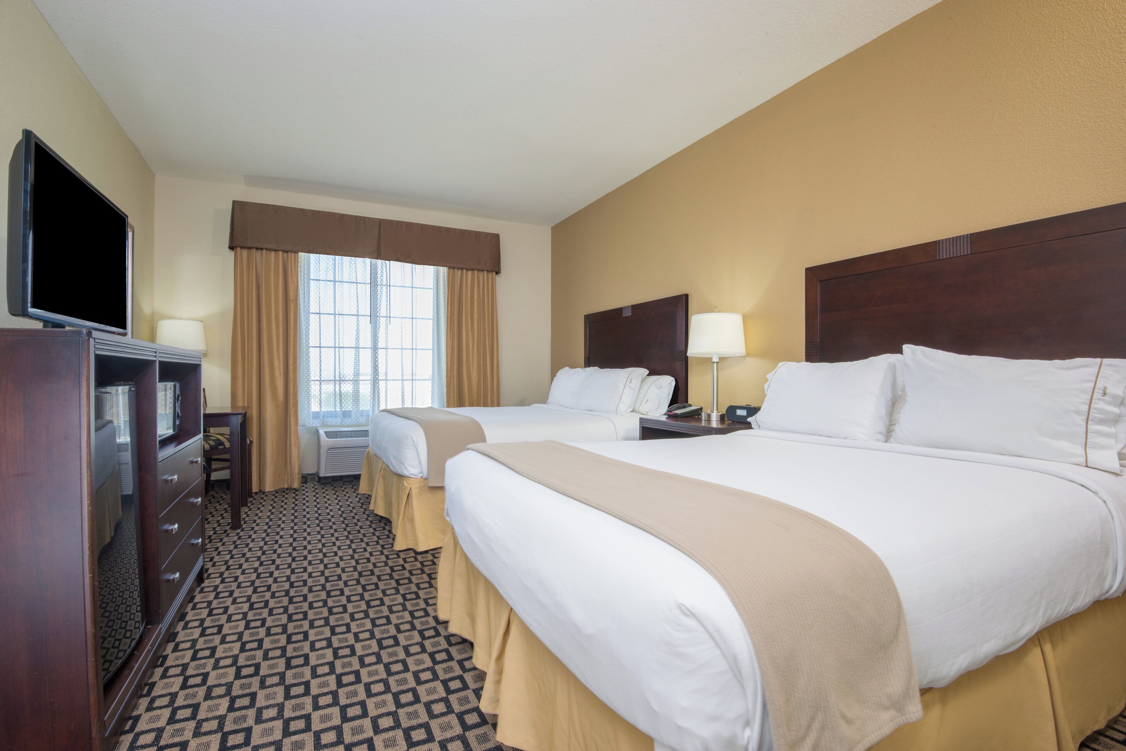 A hotel room with two beds, a TV, a desk, and a window with curtains.