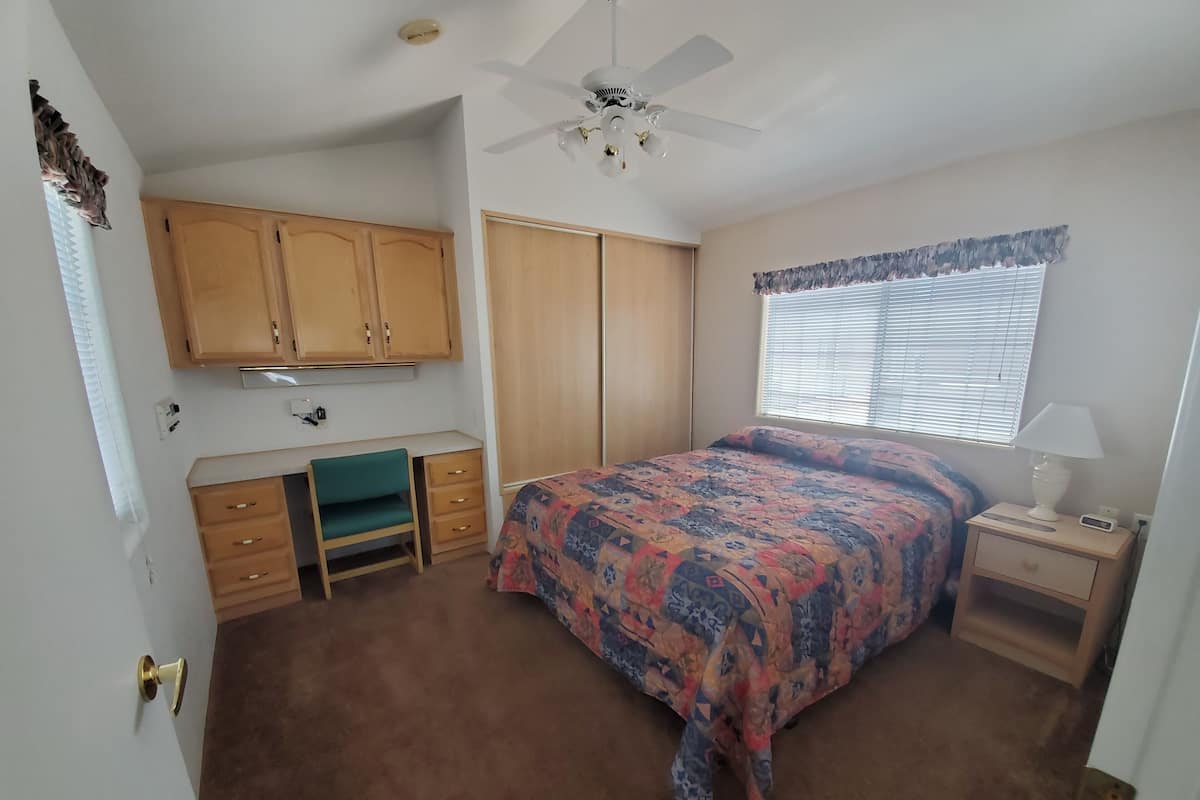 Ein Schlafzimmer mit Bett, Schreibtisch, Stuhl, Kleiderschrank, Deckenventilator und einem Fenster mit Jalousien.