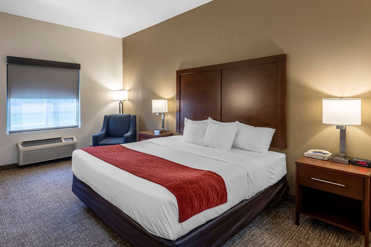 Ein Hotelzimmer mit einem großen Bett, zwei Nachttischlampen, einem Sessel, einem Heizgerät und einem Nachttisch mit einer Uhr.