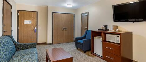 A hotel room with a sofa, a small table, a desk with a microwave, a chair, and a TV mounted on the wall.
