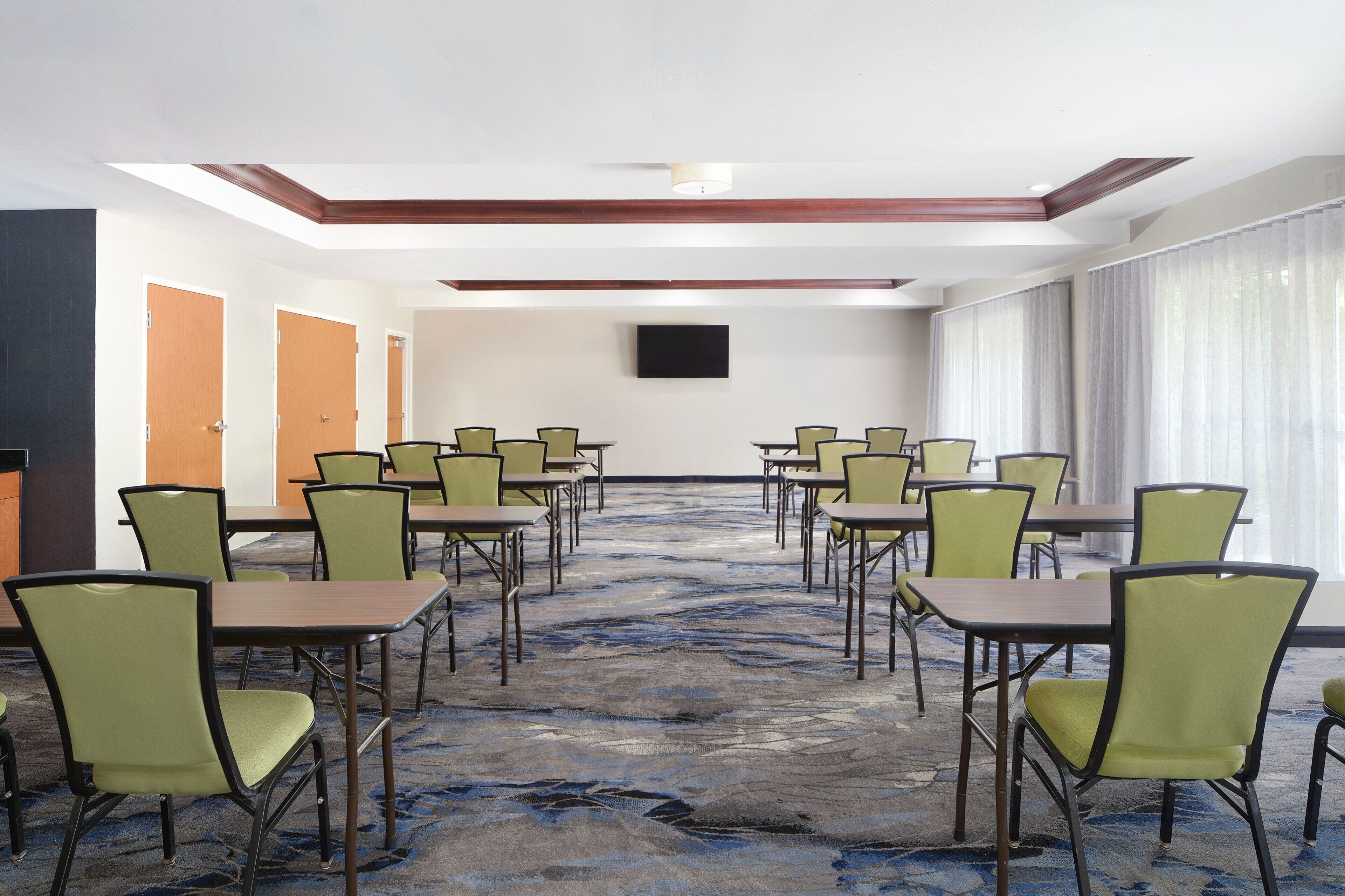 A conference room with multiple tables and chairs, a flat-screen TV mounted on the wall, and a large window with curtains.