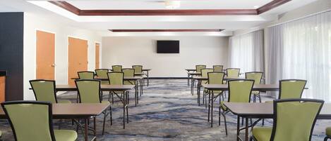 A conference room with multiple tables and chairs, a flat-screen TV mounted on the wall, and a large window with curtains.