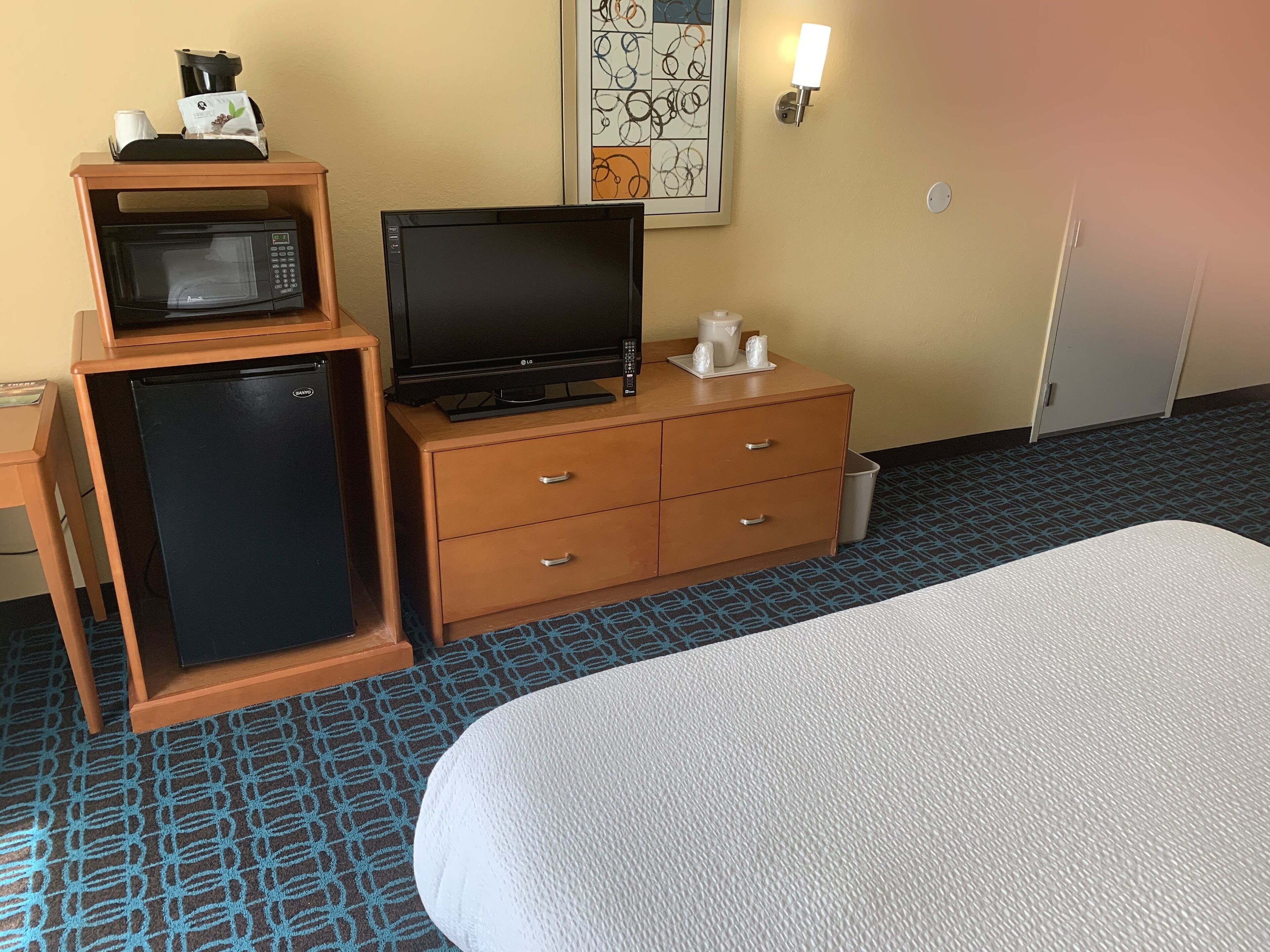 A hotel room with a bed, a minibar, a television, a coffee maker, and a wall art piece.