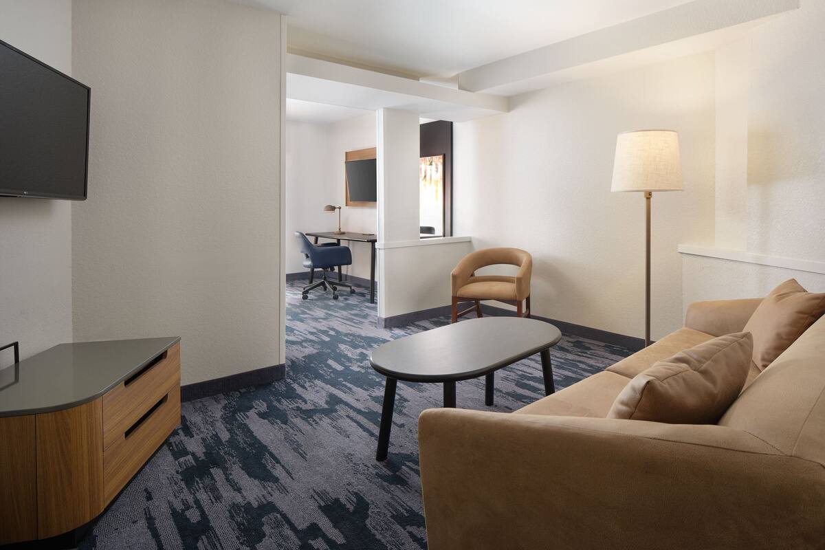 A hotel room with a sofa, a small table, a TV mounted on the wall, a desk with a chair, and a lamp.