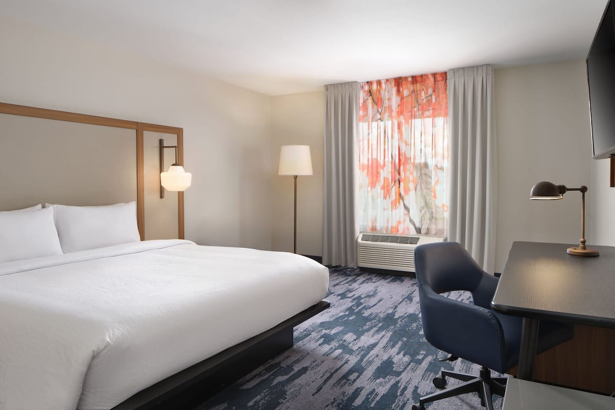 A hotel room with a large bed, a desk with a chair, a flat-screen TV, a standing lamp, and a window with curtains.